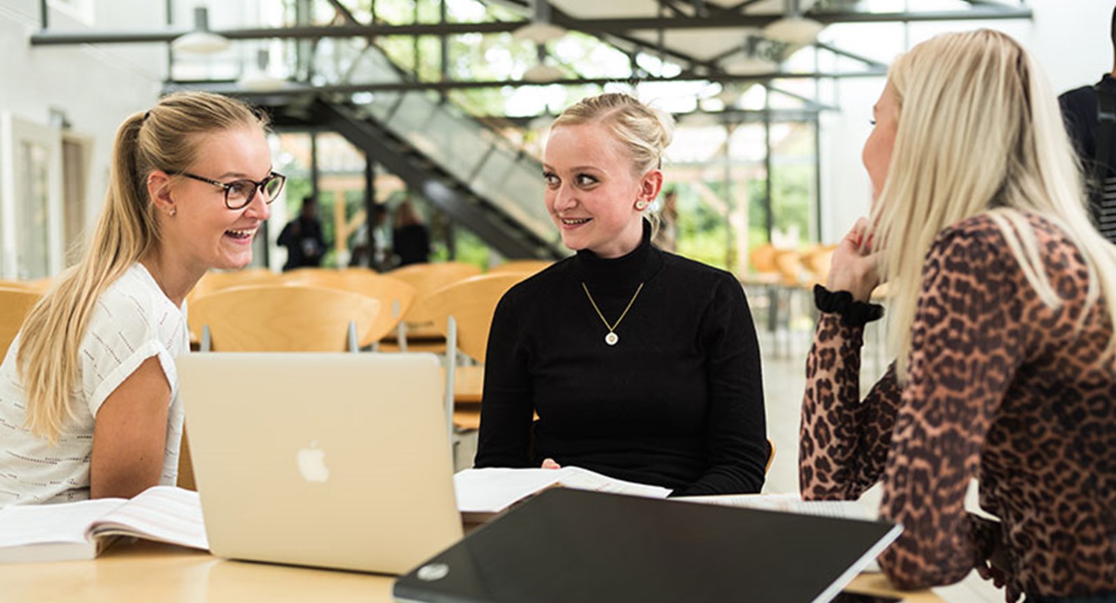 tre kvindelige studerende med computer er i samtale og smiler