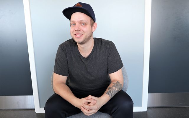 Søren i t-shirt og kasket med foldede hænder sidder foran blågrå væg