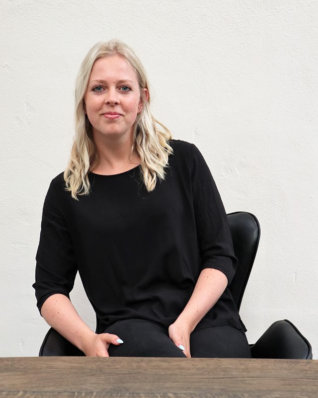 Handelsøkonom Kamilla sidder med benene over kors i en sort stol ved plankebord i sort bluse og smiler til dig