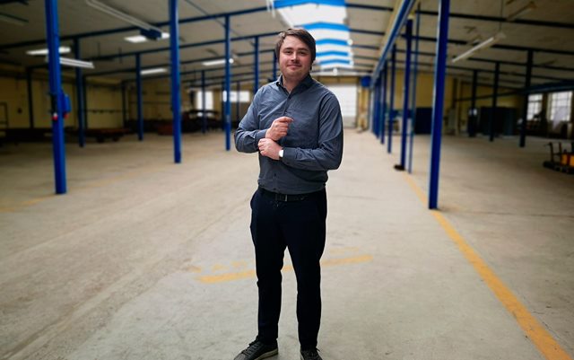 Iværksætter Kasper Reisner står midt i en stor lagerhal mellem to rækker af blå bjælker og retter på sit skjorteærme, imens han kigger ind i kameraet. 
