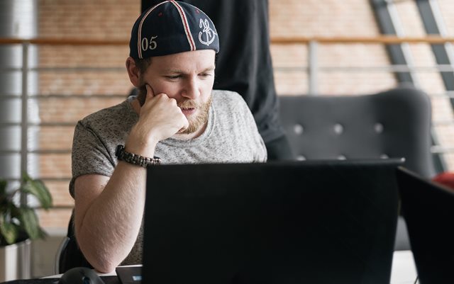 Ung mand med grå t-shirt og omvendt kasket sidder ved et bord og kigger ind i skærmen på en bærbar computer.