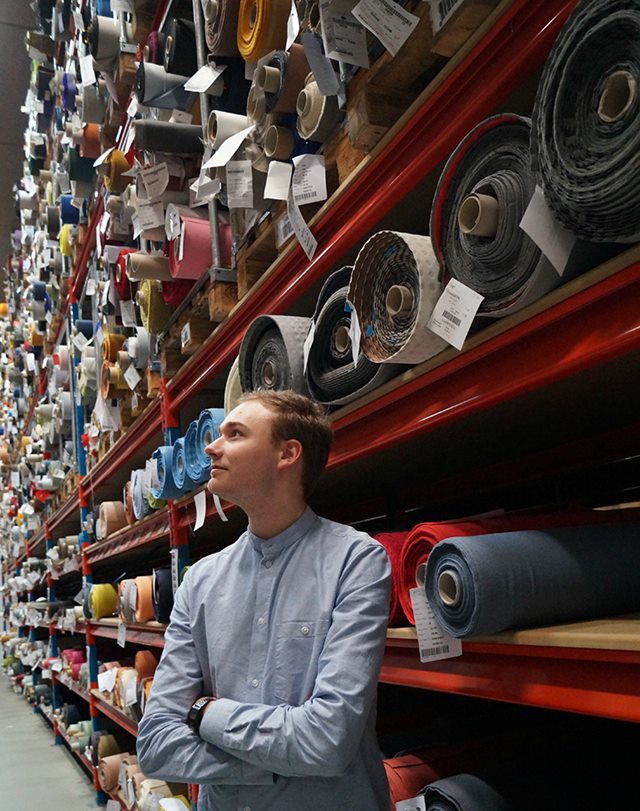 Markedsføringsøkonom-studerende Rasmus Urbak iført lyseblå skjorte står med armene over kors foran en stor industrihylde fyldt med ruller af stof i forskellige farver.