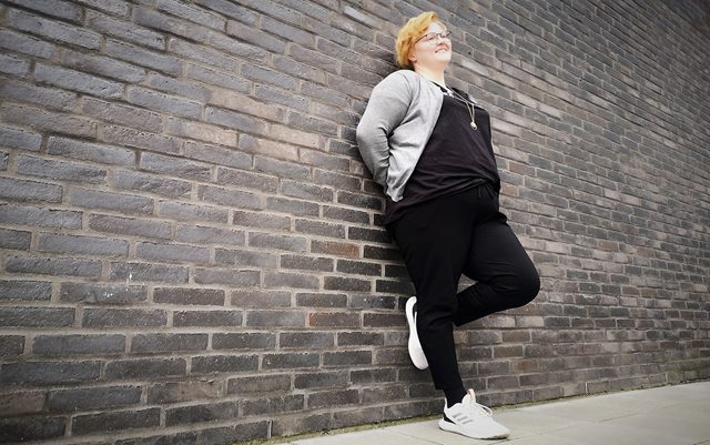 Serviceøkonom Emma Rasmussen læner sig op ad sort murstensvæg og kigger på dig