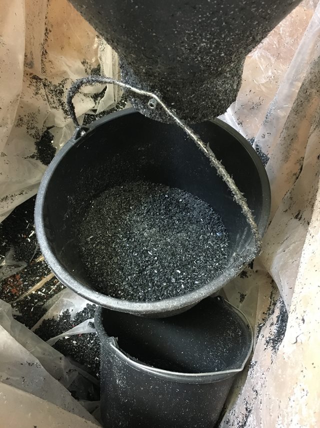 Sort malerspand hænger under en tragt og bliver fyldt med små sorte stykker af plastikgranulat.