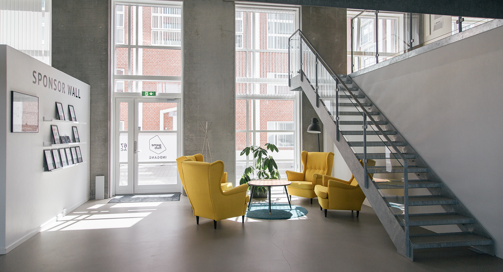 Indgangen ved Game Hub Denmark med gule lænestole om bord, grå betonvægge og trappe op på 1 sal.