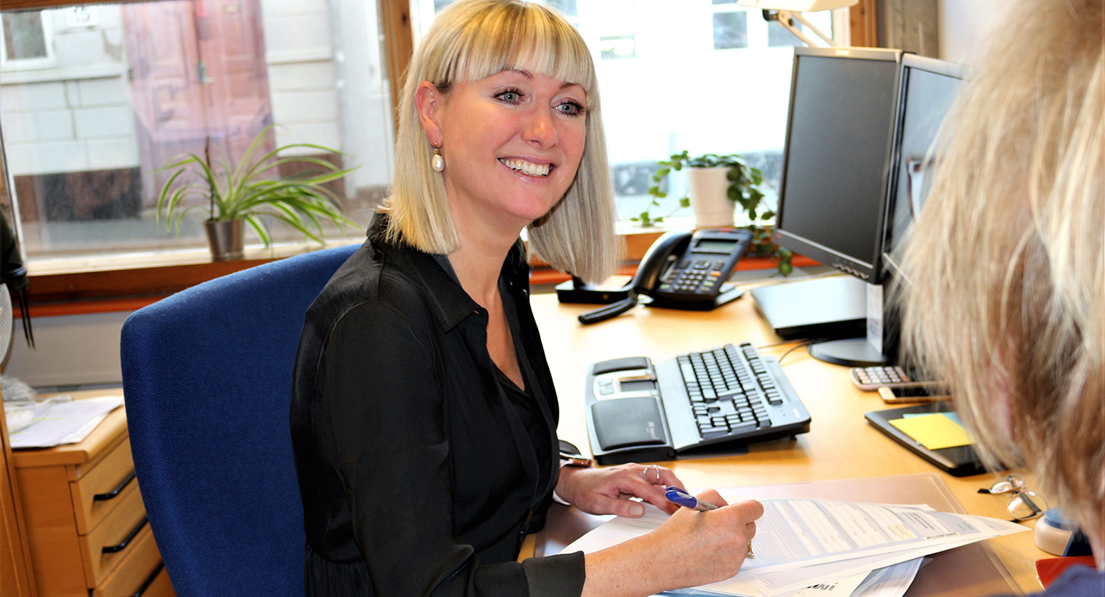 Administrationsøkonom Mette smiler til ukendt kvinde, mens hun sidder ved sit skrivebord og tager notater på papir