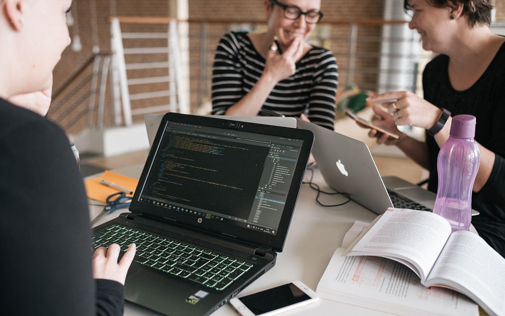 Koder på computerskærm, som datamatiker-studerende sidder ved med to kvinder i baggrunden