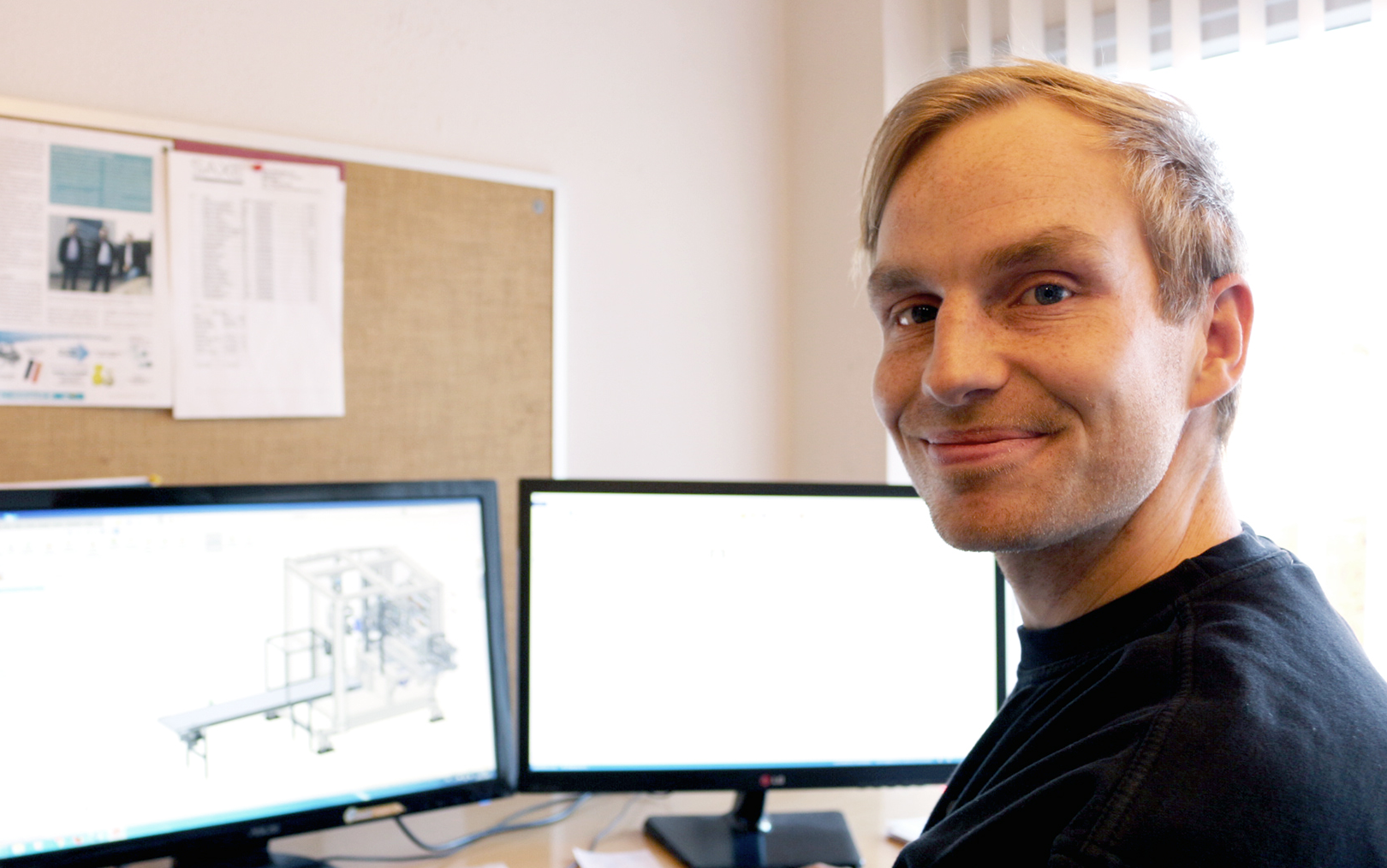 Morten smiler ved sit skrivebord med to skærme i baggrunden