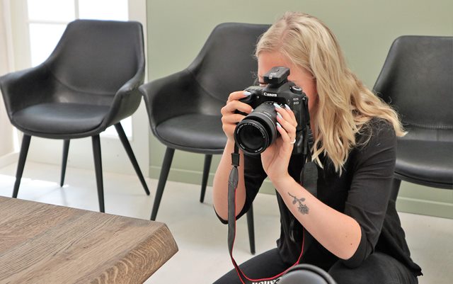 Kamilla fotograferer et bord med et stort spejlreflekskamera. I baggrunden står tre sorte stole op ad en grøn væg