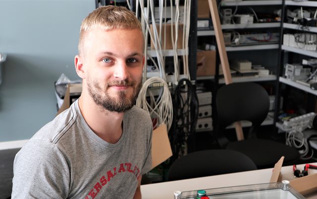 Automationsteknolog Anders Bonde smiler til dig med ledninger og PLCer i baggrunden