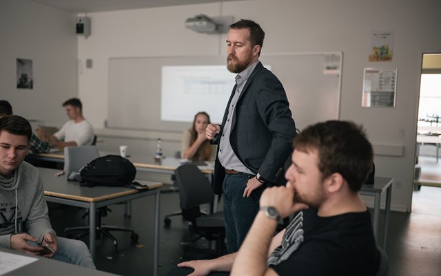 Claus Grand Bang står i et klasselokale med studerende omkring sig og kigger ned på deres papir på bord