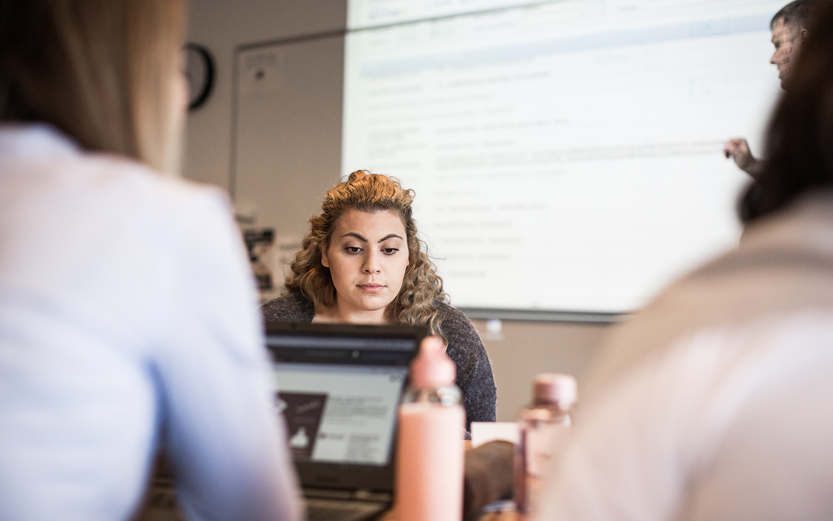 Studerende kigger ned i skræm, mens der bag hende står en person og underviser ved en tavle oplyst af en projektor