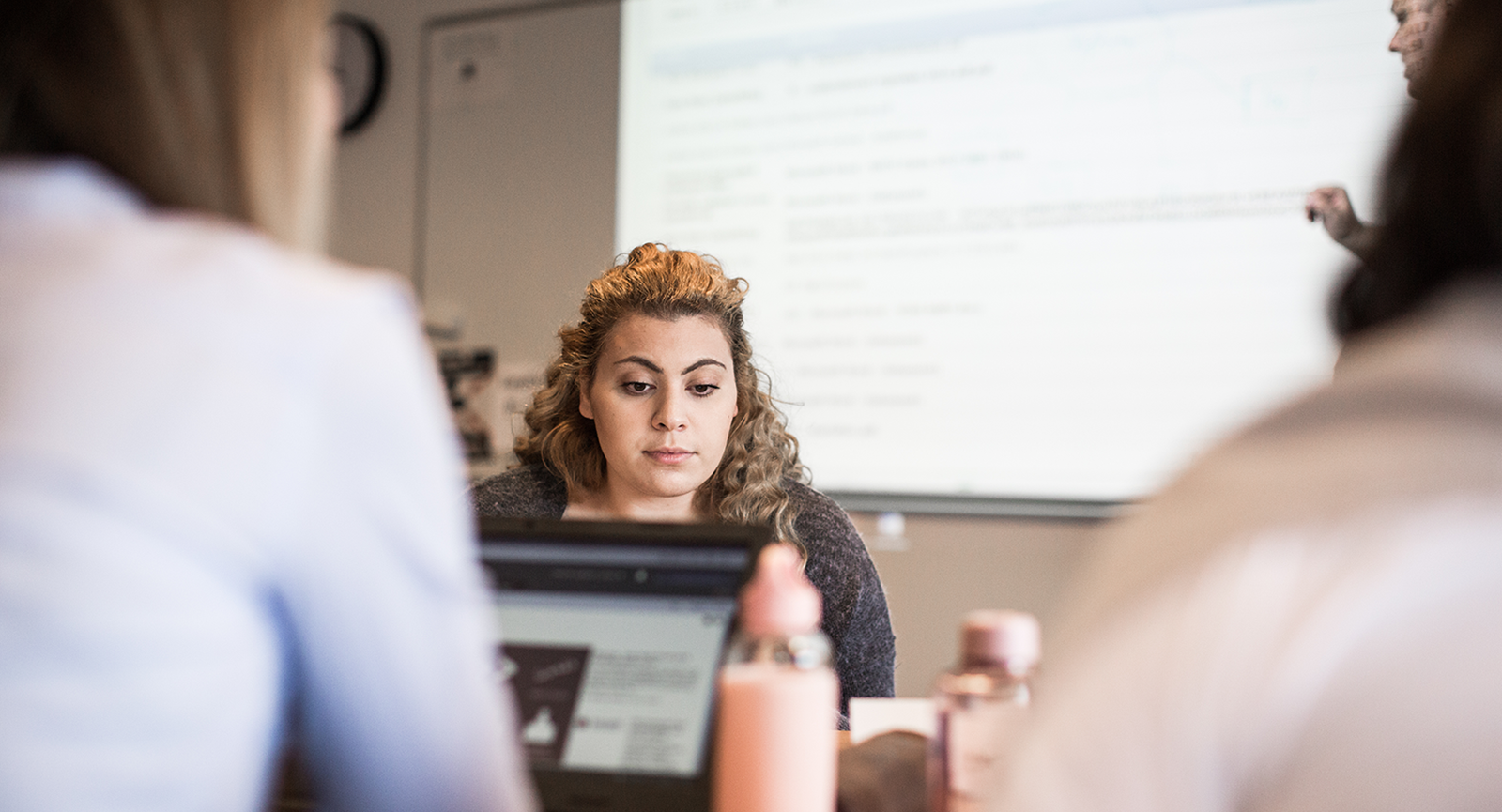 Studerende kigger ned i skræm, mens der bag hende står en person og underviser ved en tavle oplyst af en projektor