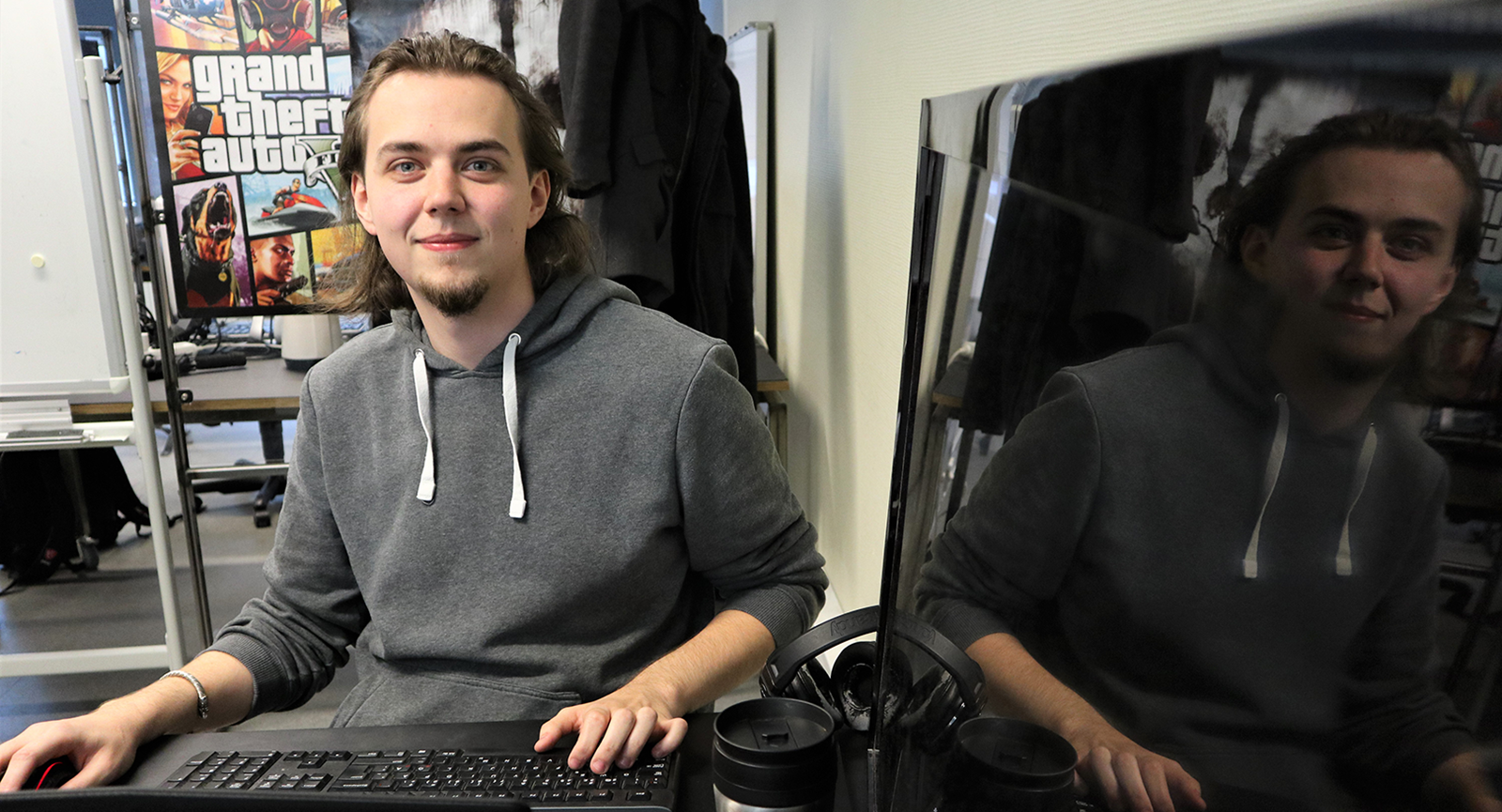 Datamatiker Levi More smiler til dig ved sit tastatur med en sort tv-skræm ved siden af sig, som viser hans spejlbillede