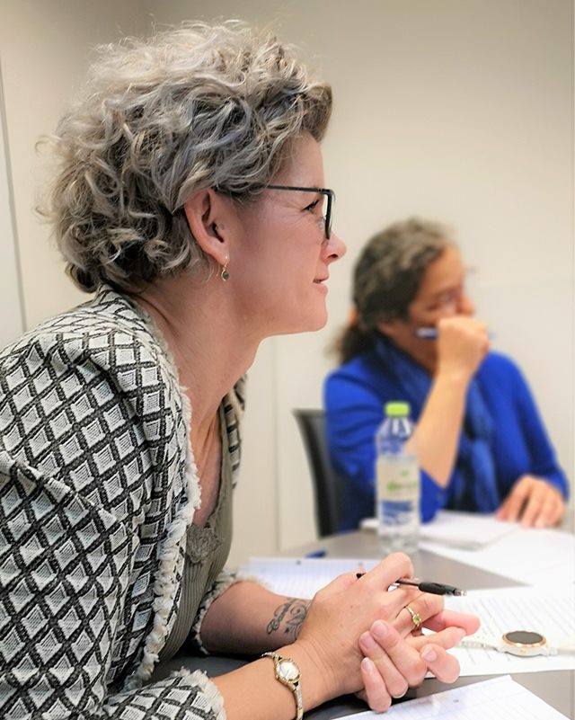 Dicte Jespersen sidder foroverbøjet ved bord med papirer foran sig og foldede hænder, mens hun lytter til en anden deltager på coaching uddannelse