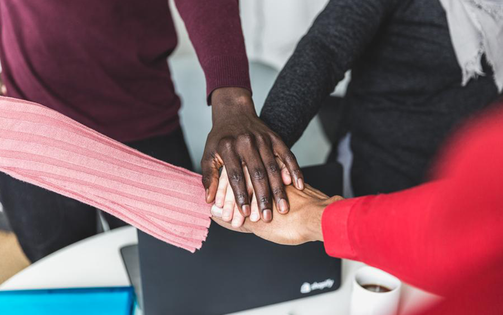 Fire forskellige hænder rører hinanden som team med kaffekop og computer i baggrunden