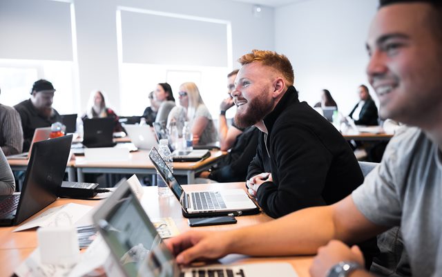 Mandlig studerende i fokus til undervisning med flere studerende tæt på, der alle smiler mod tavlen