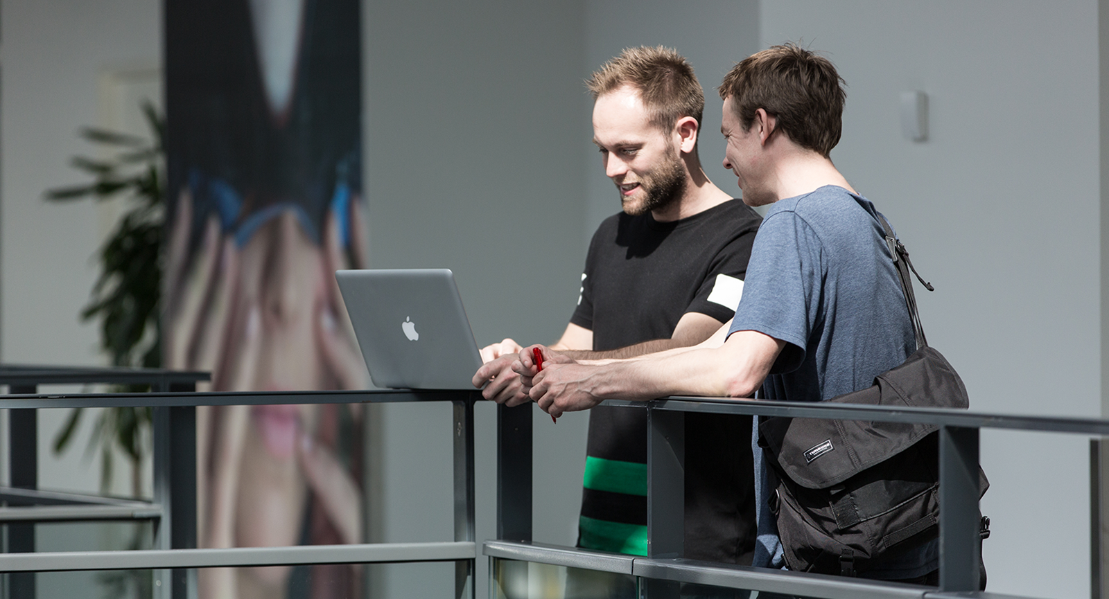 To mandlige studerende læner sig op ad et gelænder, imens de begge kigger på en bærbar computer.