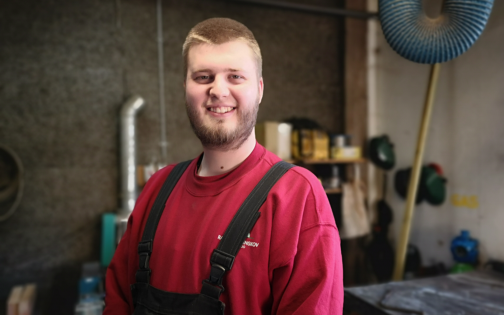 Piet Mortensen står på et værksted og smiler til kameraet iført en rød bluse og mørkeblå overalls. 