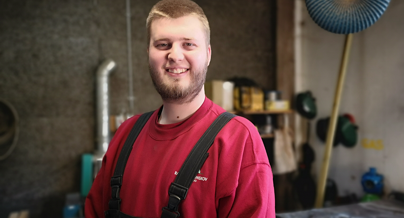 Piet Mortensen står på et værksted og smiler til kameraet iført en rød bluse og mørkeblå overalls. 