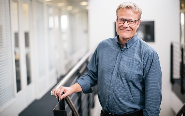 Rektor Anders Graae Rasmussen står ved en trappe med den ene hånd på gelænderet og smiler til kameraet. 