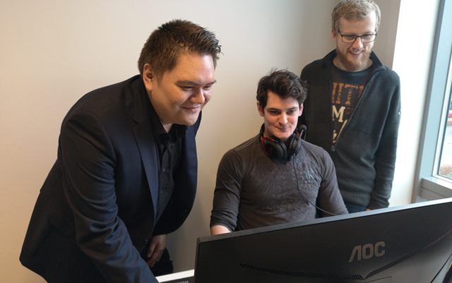 Tre mænd kigger interesseret på den samme computerskærm. Manden i midten sidder på en stol, imens de to andre mænd står på hver sin side af ham.