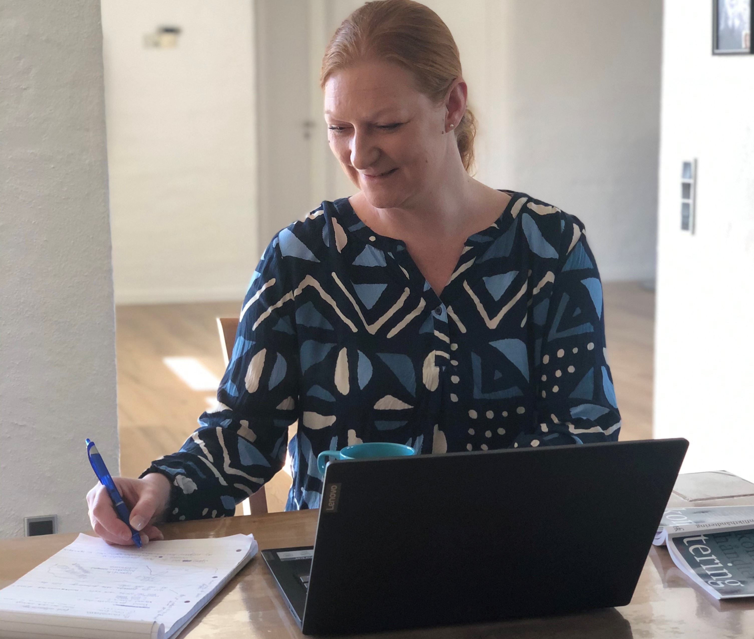 Socialrådgiver Diana Laursen tager noter på en notesblok, mens hun sidder ved et spisebord foran en bærbar computer og holder om en lyseblå kaffekop.