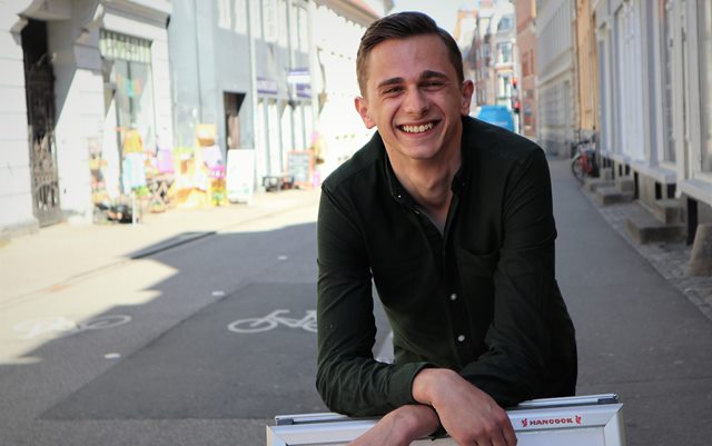 Markedsføringsøkonomstuderende Marko Bubalo står midt på Mejlgade i Aarhus og læner sig op ad et skilt, mens han storsmiler ind i kameraet.