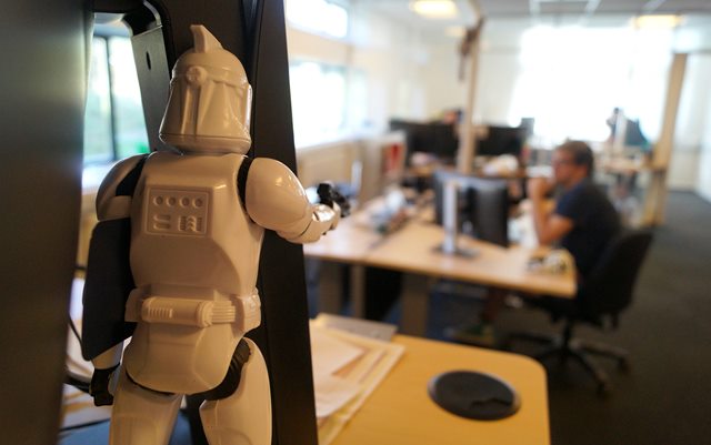 Nærbillede af en hvid actionfigur fra Star Wars, som står med ryggen til kameraet på et skrivebord i et kontor med flere skriveborde i baggrunden. 