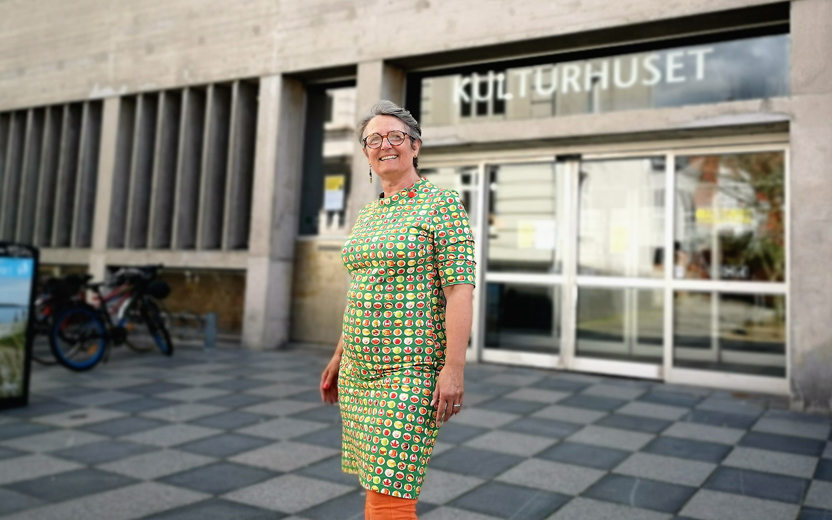Bibliotekar Dorthe Meyer Andreassen står uden foran Randers Bibliotek og storsmiler iført en grøn kjole med en masse prikker i gule og røde farver.