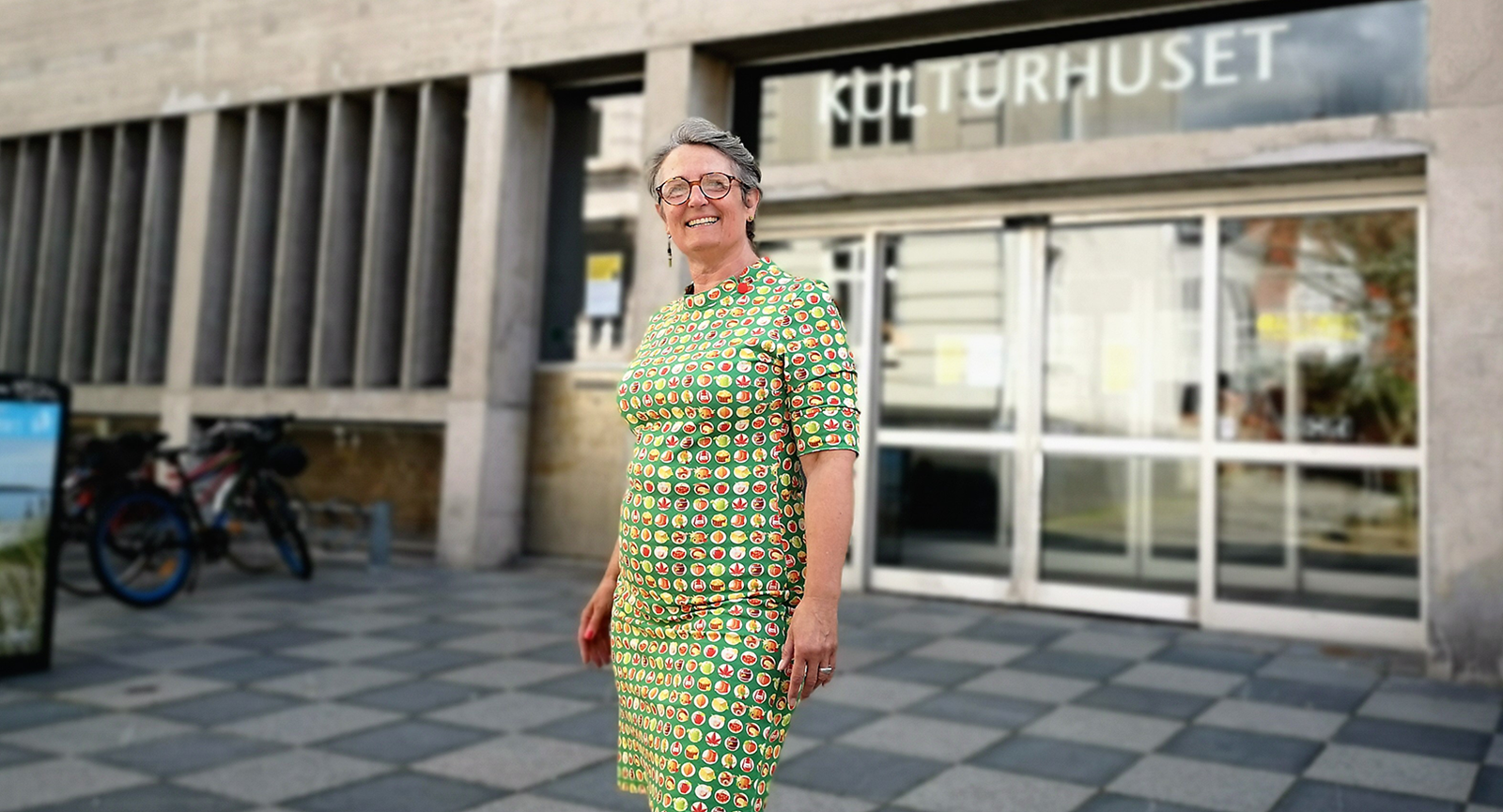 Bibliotekar Dorthe Meyer Andreassen står uden foran Randers Bibliotek og storsmiler iført en grøn kjole med en masse prikker i gule og røde farver.