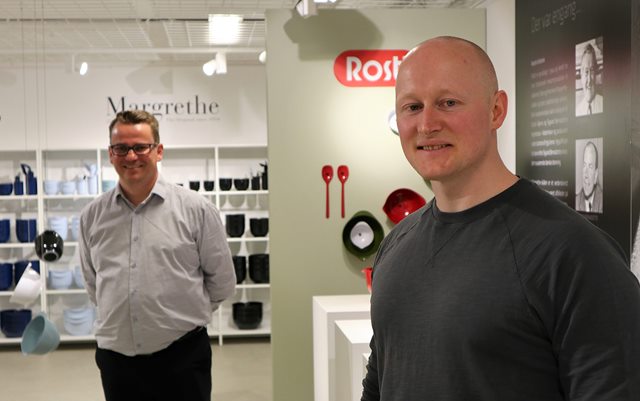 Logistikøkonom-studerende Dennis Mortensen står i et showroom fyldt med køkkengrej hos virksomheden F&H og smiler til kameraet. I baggrunden står F&H's logistikdirektør Ruddi Vestergaard Nielsen.