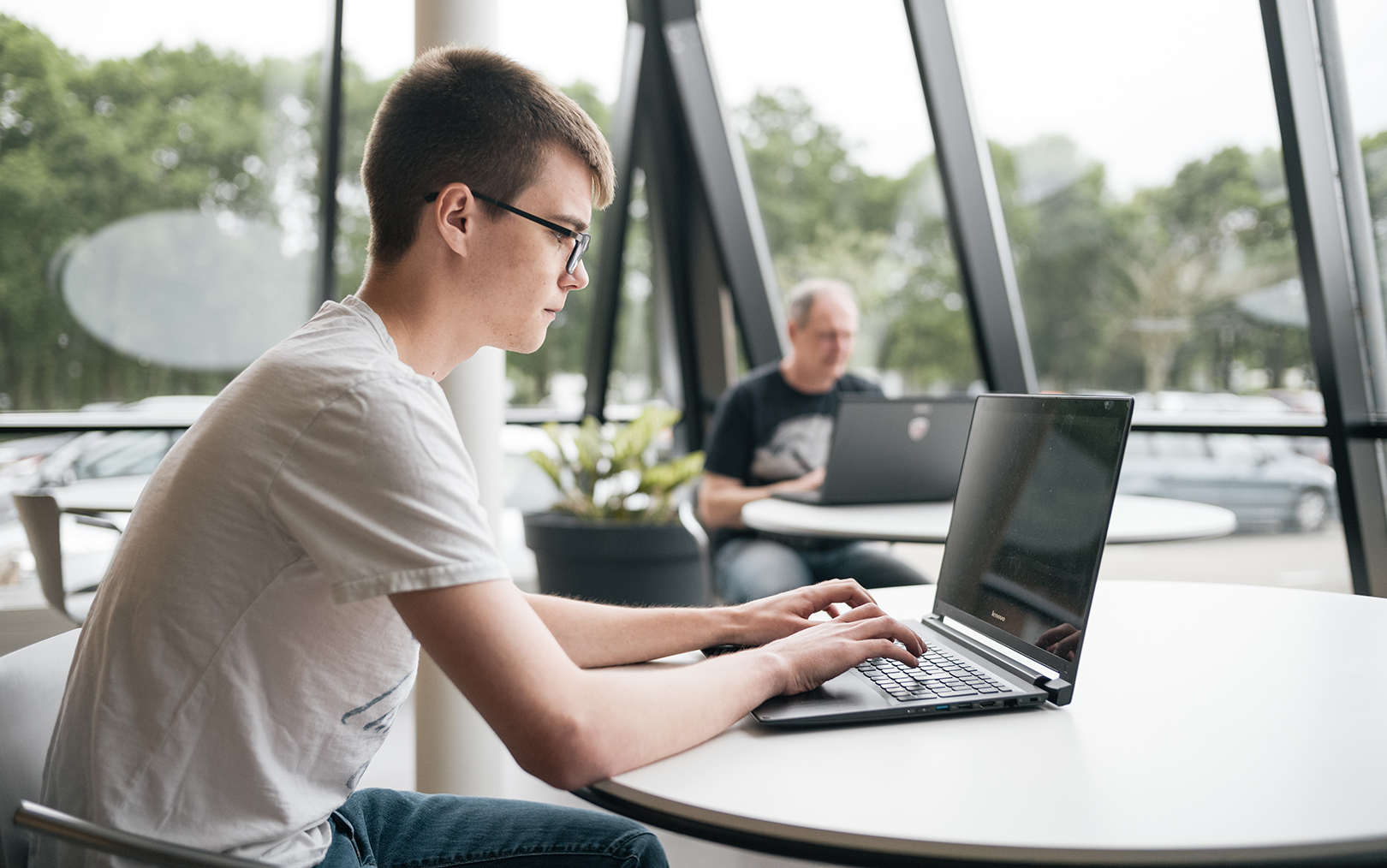 Ung mand i hvid t-shirt og mørke briller sidder ved et bord og arbejder koncentreret på en bærbar computer.