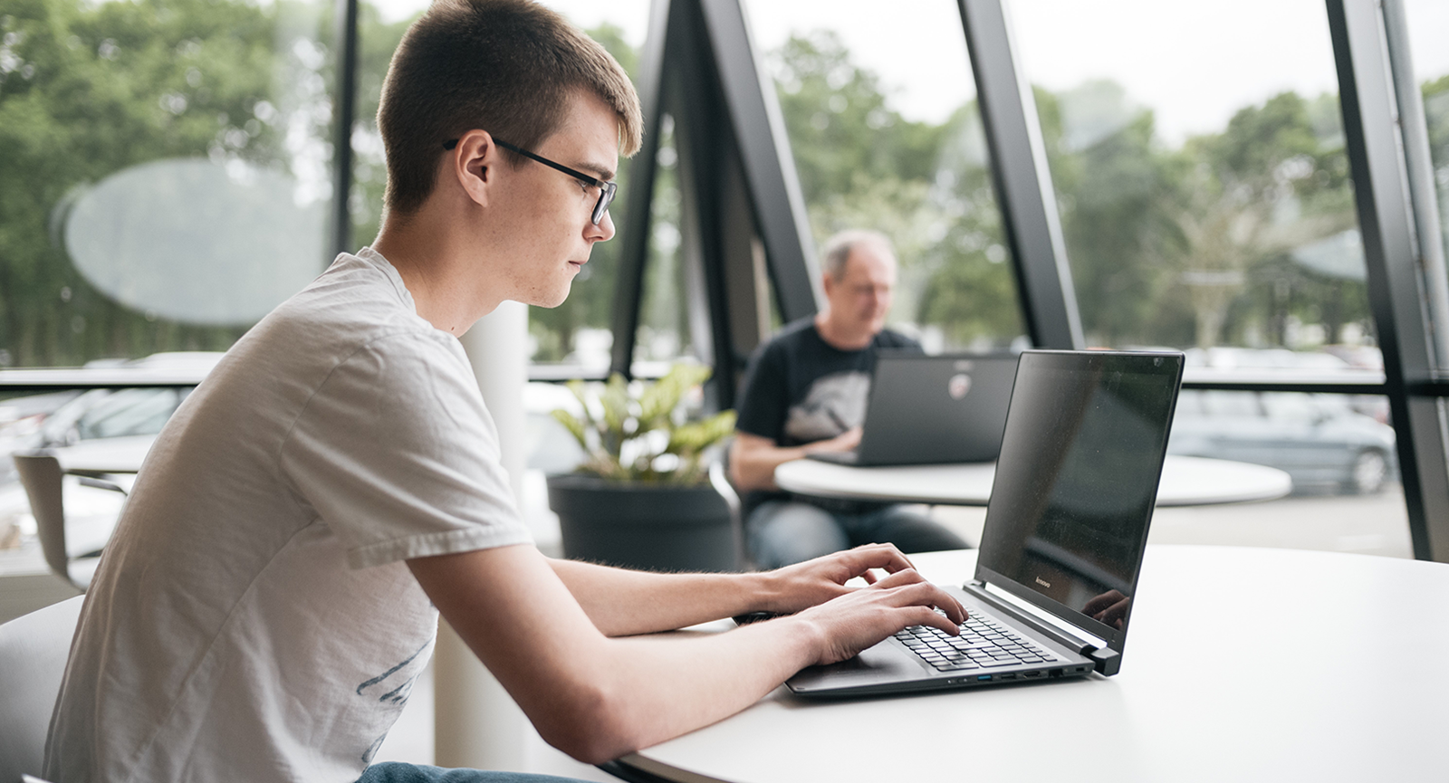 Ung mand i hvid t-shirt og mørke briller sidder ved et bord og arbejder koncentreret på en bærbar computer.