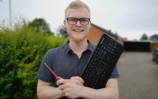 Nyuddannet automationsteknolog Rasmus Stampe Laursen står i en indkørsel og smiler til kameraet med en rød skruetrækker i den ene hånd og et sort tastatur i den anden.