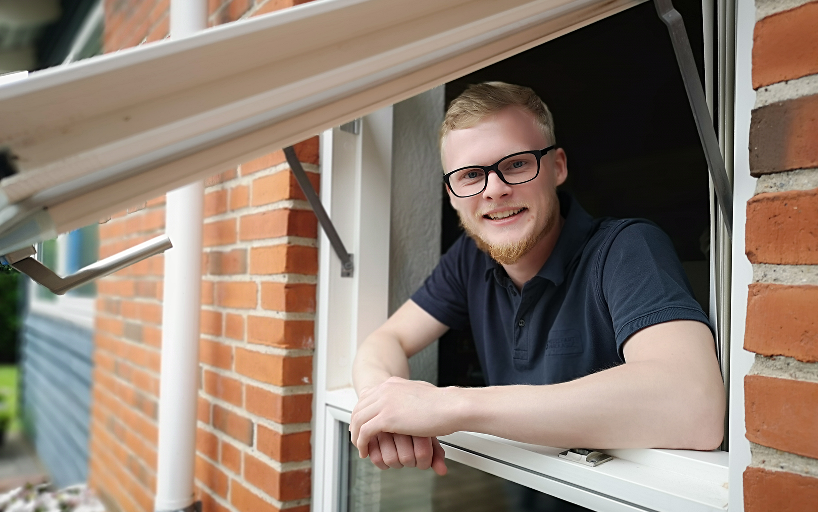 Nyuddannet automationsteknolog Rasmus Stampe Laursen læner sig ud ad vinduet på et rødt murstenshus, mens han hviler albuerne på vindueskarmen og smiler til kameraet. 