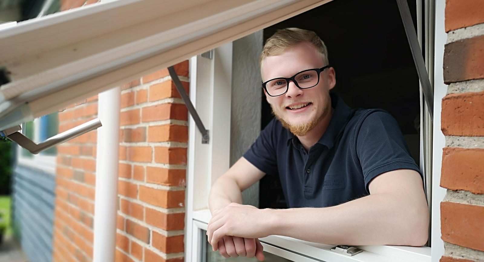 Nyuddannet automationsteknolog Rasmus Stampe Laursen læner sig ud ad vinduet på et rødt murstenshus, mens han hviler albuerne på vindueskarmen og smiler til kameraet. 
