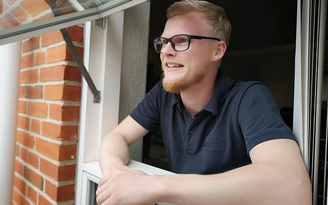 Nyuddannet automationsteknolog Rasmus Stampe Laursen læner sig ud ad vinduet på et rødt murstenshus, mens han hviler albuerne på vindueskarmen og kigger ud mod husets have.
