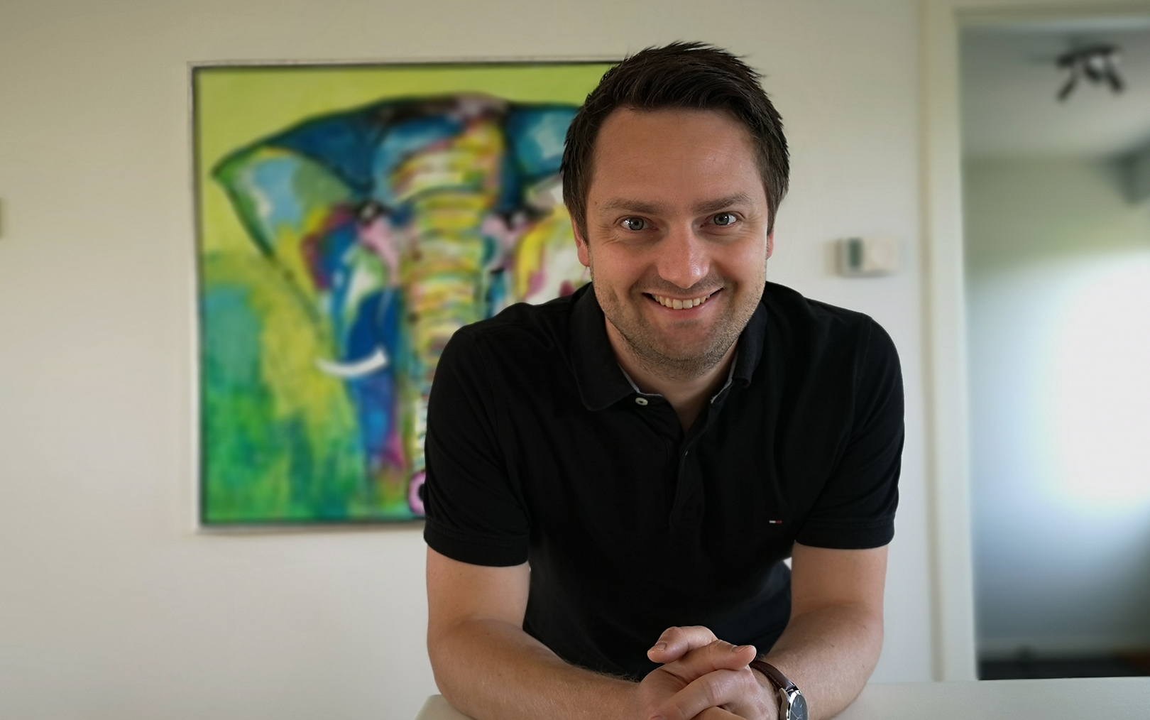 Portrætfoto af den tidligere professionelle pokerspiller Morten Christensen, der har søgt ind på uddannelsen i dataanalyse. Han smiler til kameraet, imens han hviler albuerne på et bord. I baggrunden ses et maleri af en elefant i gule og grønne nuancer.