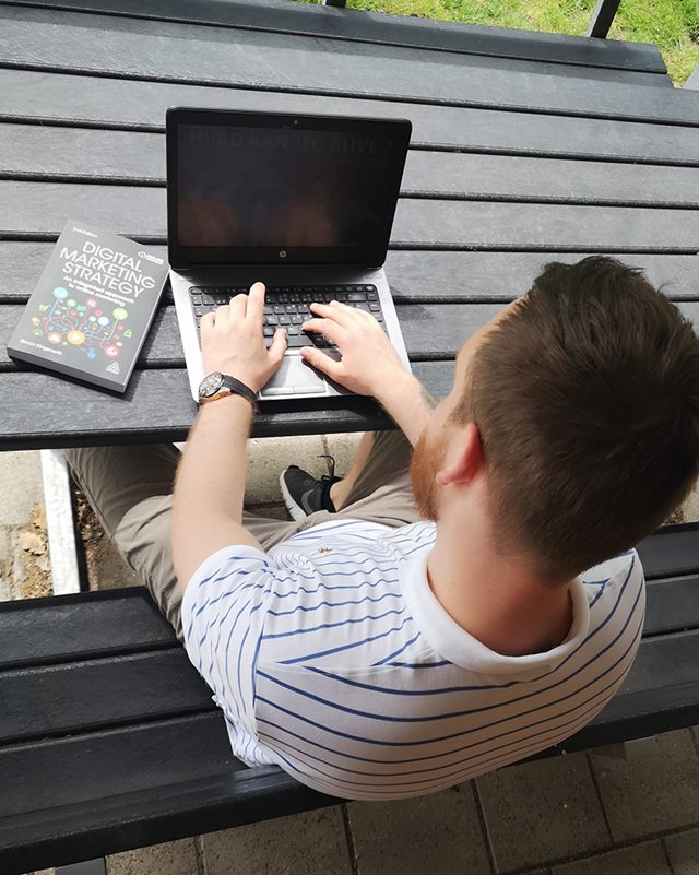 René Klokkerholm sidder udenfor ved et bord-bænkesæt og arbejder på en bærbar computer. Billedet er taget oppefra. Han er iført en blåstribet polo t-shirt. På bordet ved siden af computeren ligger en fagbog med titlen 'Digital Marketing Strategy'.