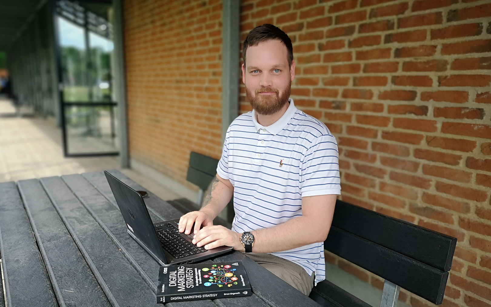 René Klokkerholm sidder udenfor ved et bord-bænkesæt og arbejder på en bærbar computer, mens han kigger til siden direkte mod kameraet.