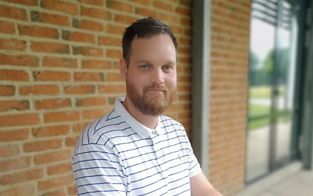 Portrætfoto af René Klokkerholm, der sidder ved et bord-bænkesæt med en rød murstensmur i baggrunden og kigger lige mod kameraet iført en hvid og blåstribet polo t-shirt.