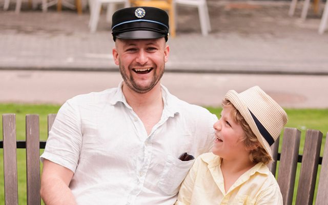 En mand og en dreng sidder udenfor på en bænk. Manden er iført kortærmet, hvid skjorte og sort studenterhue, og han storsmiler direkte mod kameraet. Drengen er iført lys skjorte og sivhat og ser smilende op på manden.