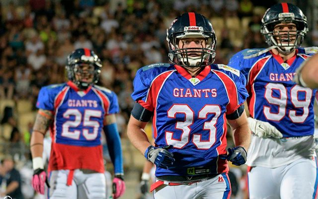 Tre NFL-spillere i fuld spillerdragt på banen med en fyldt tilskuertribune i baggrunden. Spillerne bærer blå spillertrøjer fra den amerikanske fodboldklub New York Giants samt hjelme, handler og hvide bukser.