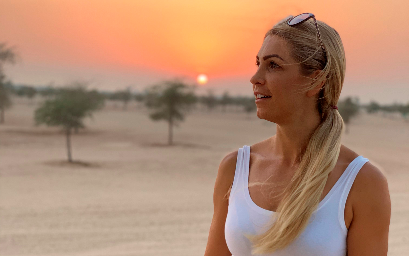 Personlig træner Tina Tougaard står med siden til kameraet i en ørken med nogle få små træer. Solnedgangen farver himlen orange. Hun er iført hvid top og solbriller i panden.