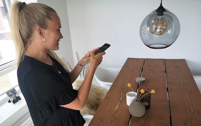 Handelsøkonom Cecilie Blaaberg står ved et lille spisebord med sin telefon i hænderne i gang med at tage et billede af bordet. På bordet ses nogle lysestager til fyrfadslys og en lerkrukke med tre gule blomster i.