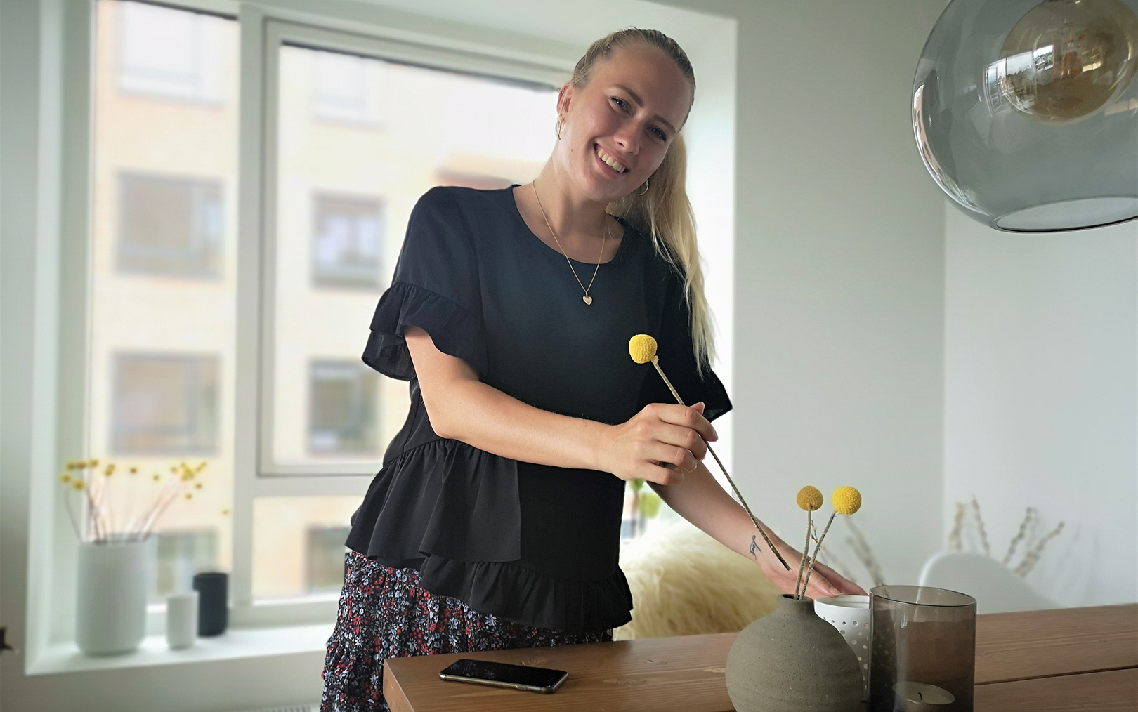Handelsøkonom Cecilie Blaaberg står bag et spisebord i en lejlighed og smiler mod kameraet med ryggen til et vindue. Hun er i gang med at anrette tre gule blomster i en rund lerkrukke på bordet.