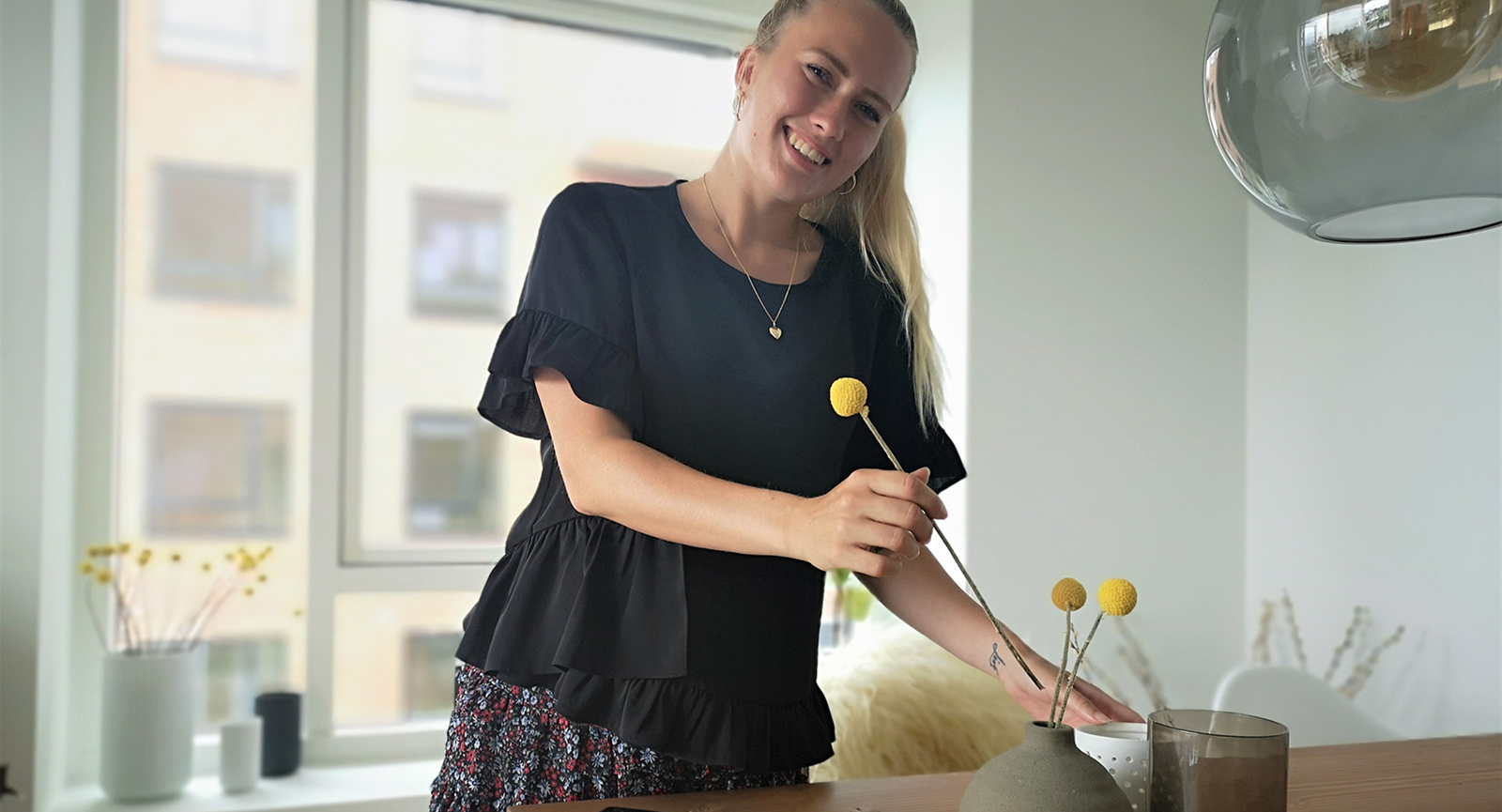 Handelsøkonom Cecilie Blaaberg står bag et spisebord i en lejlighed og smiler mod kameraet med ryggen til et vindue. Hun er i gang med at anrette tre gule blomster i en rund lerkrukke på bordet.