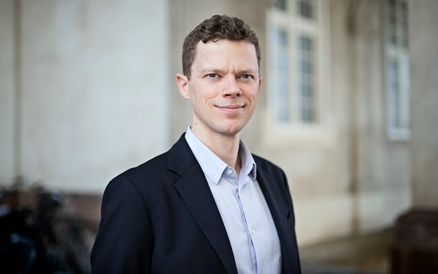 Portrætfoto af Mads Eriksen, som er uddannelses- og forskningspolitisk chef ved Dansk Erhverv. Han er iført lyseblå skjorte og mørk jakke og kigger smilende mod kameraet, mens han står foran en lysegrå bygning.