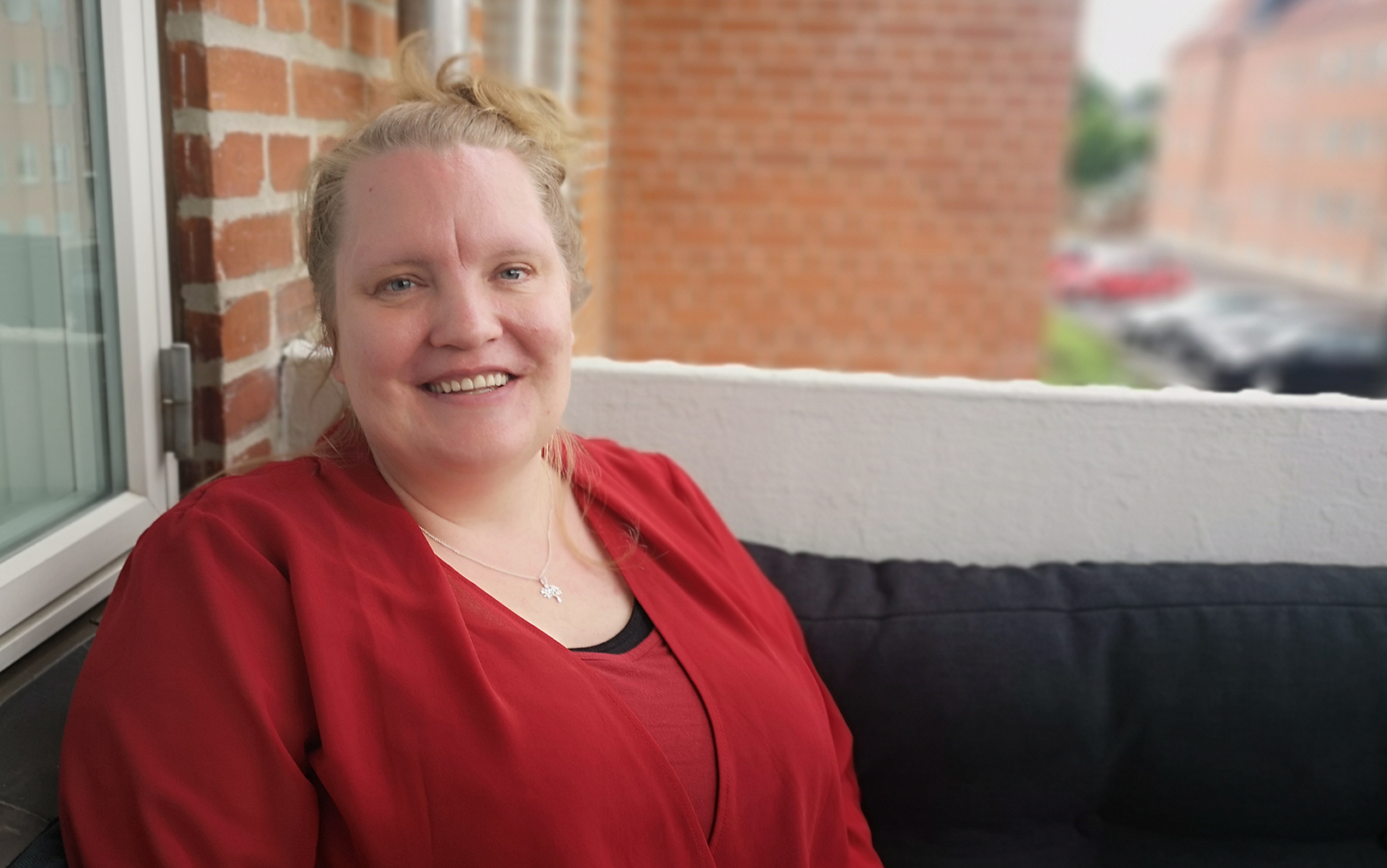 Portrætbillede af social- og sundhedshjælper Tanja Rasmussen, der sidder på en sofa på en altan i en rød murstensbygning og smiler til kameraet.